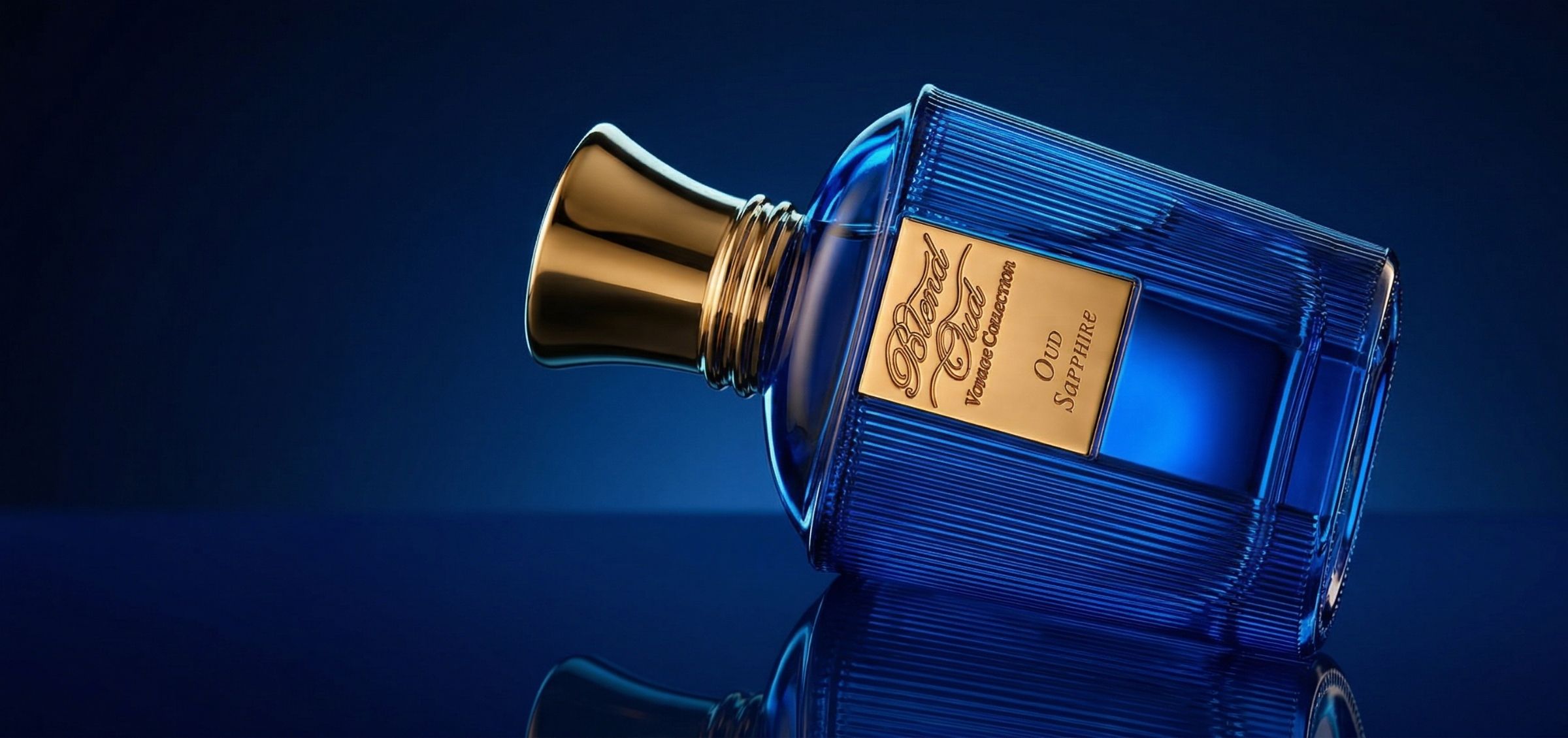 Blue glass perfume bottle tipped on its side with a gold cap, resting on a dark reflective surface.