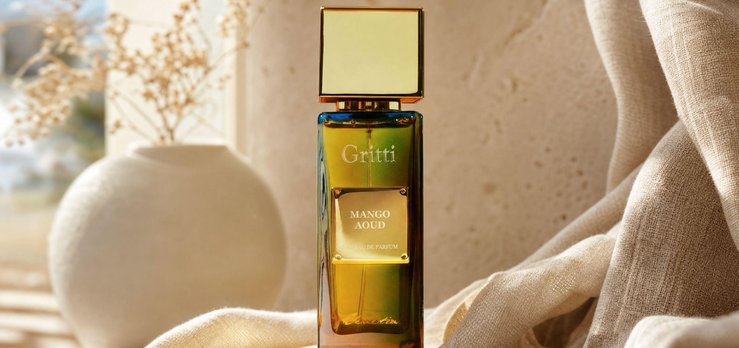 Gritti Mango Oud perfume bottle with a gold cap, amber-green glass, set on beige fabric backdrop.