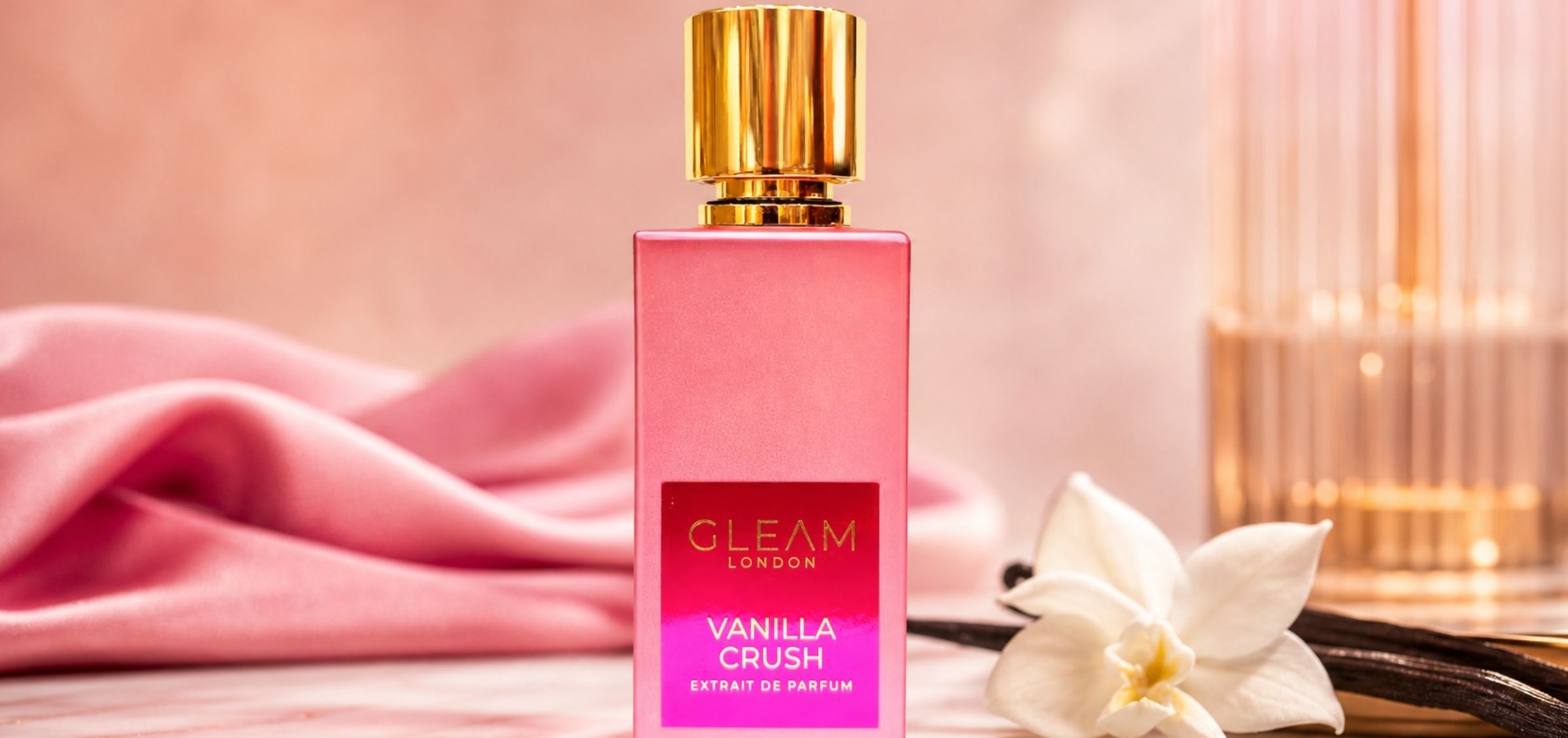 Pink perfume bottle with a gold cap, labeled 'Gleam London Vanilla Crush Extrait de Parfum', set on pink fabric.